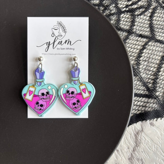 Heart-shaped earrings with skull design on a Glam by Sam Whiting card, displayed on a black surface.