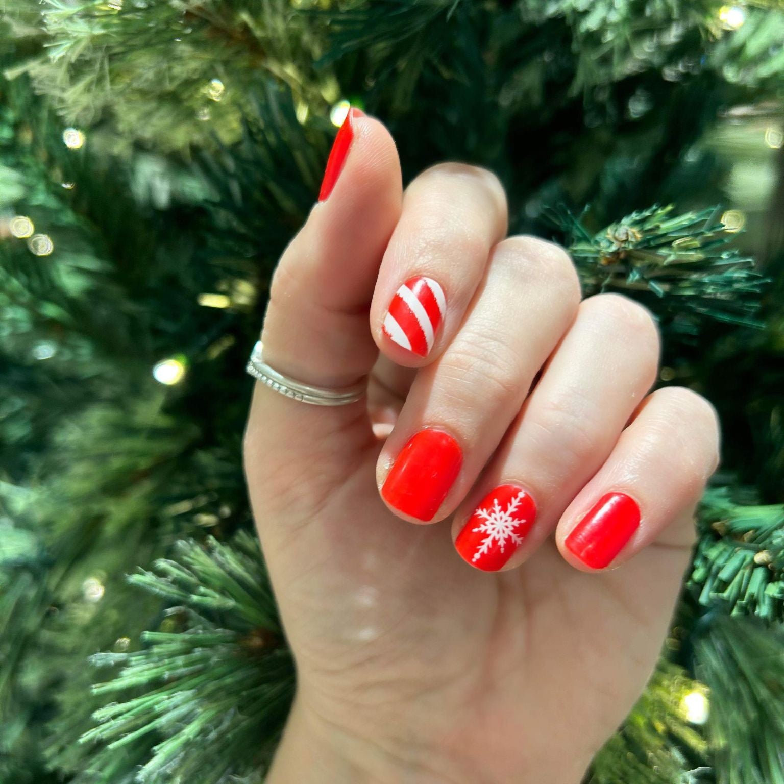 Hand with red nail polish and a snowflake design against a green Christmas tree background