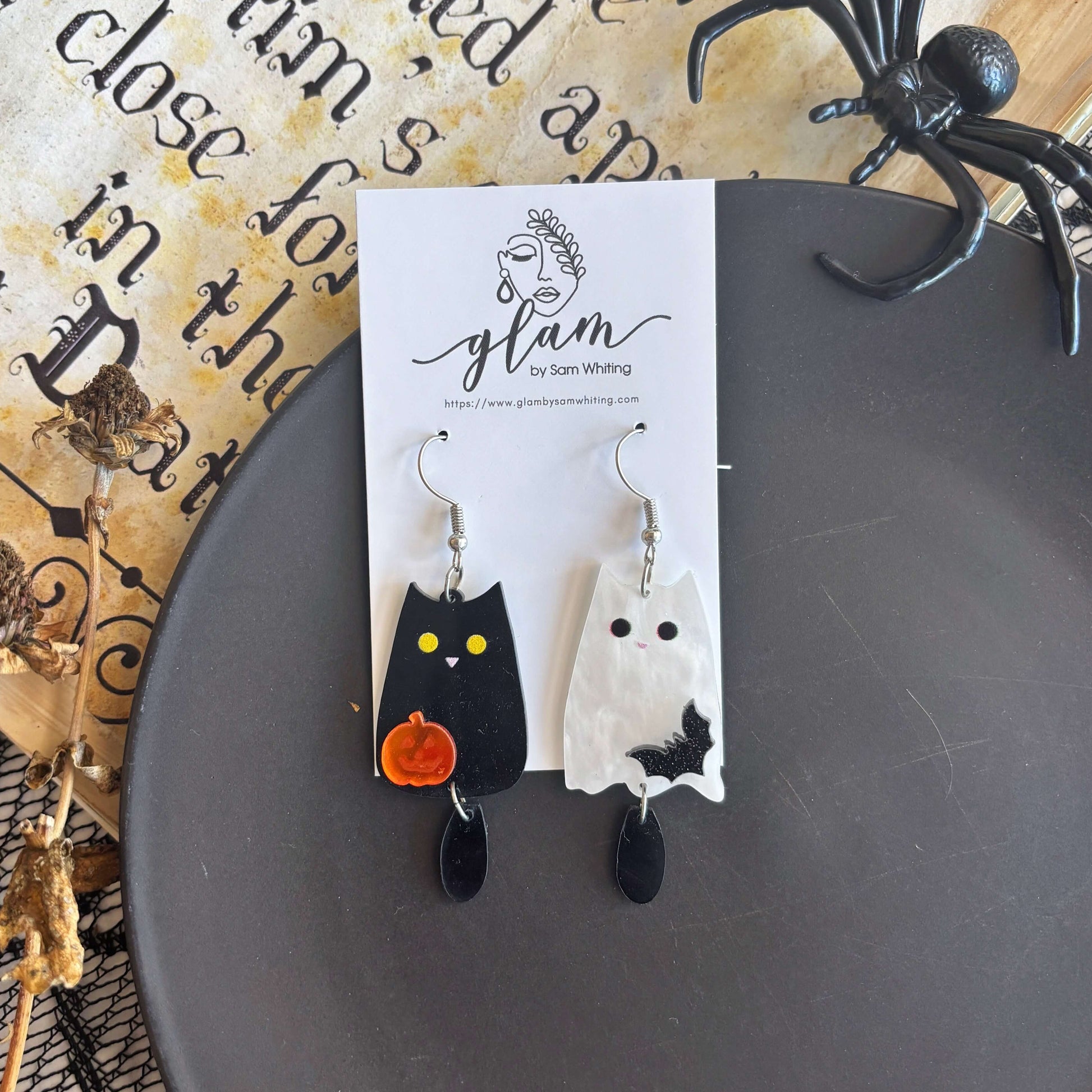 A pair of Halloween-themed dangling earrings displayed on a white card. One earring is shaped like a black cat with yellow eyes and a small orange pumpkin, and the other is a white ghost with black details.