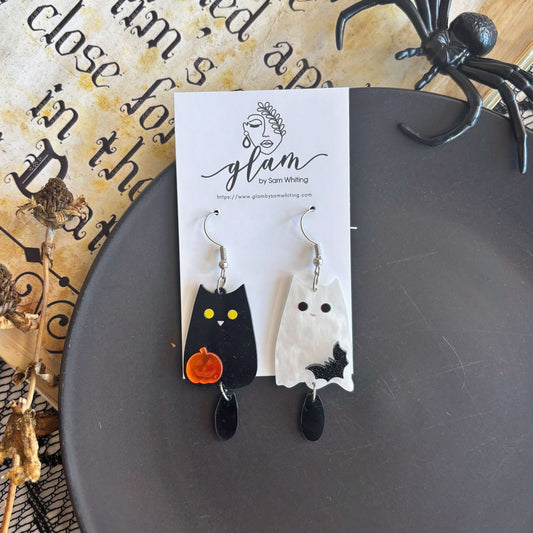 A pair of Halloween-themed dangling earrings displayed on a white card. One earring is shaped like a black cat with yellow eyes and a small orange pumpkin, and the other is a white ghost with black details.